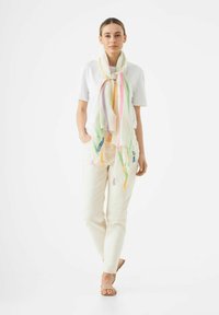Écharpe légère multicolore avec des motifs en chevron, à bords frangés, arborant des accents en rose pastel, vert et jaune. Portée sur une chemise blanche.