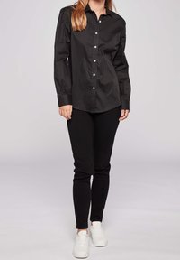 Černá košile na knoflíky s dlouhými rukávy a nabíranými detaily, v kombinaci s černými kalhotami a bílými teniskami. Hladká, ležérní tkanina.