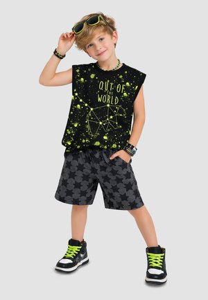 Junge trägt ein schwarzes ärmelloses Shirt mit neonfarbenem Sternbild eines Bären und dem Schriftzug „Out of the World“, sternenbedruckte Shorts und Sneakers mit neonfarbenen Schnürsenkeln.