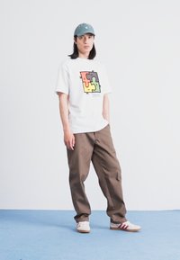 Weißes T-Shirt mit buntem Puzzle-Motiv; braune lockere Hose; blauer Hut; weiße Sneakers mit roten Streifen; einfarbiger Hintergrund.
