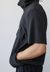 Camisa negra de manga corta con cuello alto y bolsillo plano en el frente. Textura suave y mate, corte holgado y diseño minimalista.