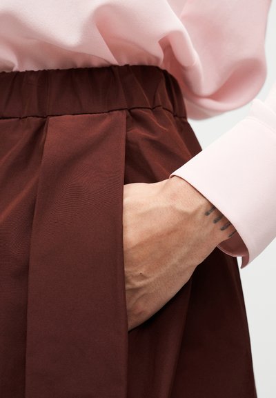 Καφέ πλισέ φούστα με ελαστική μέση· το χέρι ακουμπά σε τσέπη· απαλή υφή· συνδυάζεται με ανοιχτό ροζ μπλούζα μακρυμάνικη.