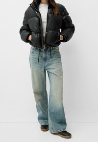 Svart uppblåsbar jacka med tryckknappar, med en kortad design och dragsko. Matchad med ljusblå vida jeans och svarta skor.