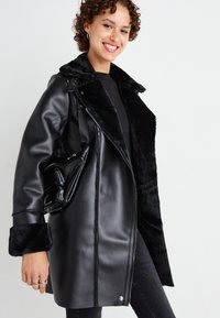 Chaqueta de cuero sintético negra con un suave collar y puños de piel negra, con cierre de cremallera y un bolsillo lateral.