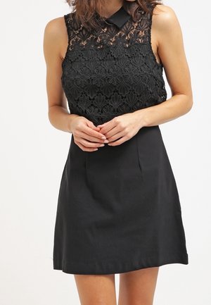 Frau trägt ein ärmelloses schwarzes Kleid mit floraler Spitzen-Steil und einem einfarbigen Rock, die Hände vor der Taille gefaltet, vor einem schlichten Hintergrund.