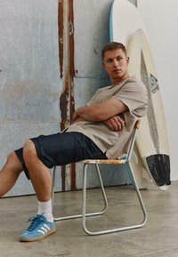 Jeune homme en chemise beige et short noir assis sur une chaise en métal, les bras croisés, à côté de deux planches de surf contre un mur usé.