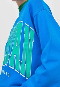 Sudadera azul con texto verde, con un escote redondeado y un corte holgado. El material parece suave y similar al algodón, con costuras visibles.