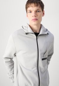 Light gray zip-up sweatshirt with a hood, featuring a front pocket, black zipper, and subtle texture. Casual and functional design.