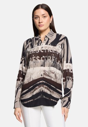 Long-sleeve button-up shirt in beige, brown, and black with abstract patterns and text. Smooth fabric with a slightly loose fit and curved hem.
