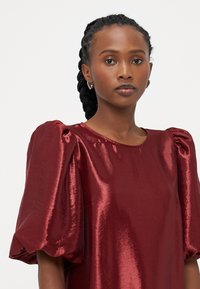 Βουργουνδί, μεταλλική μπλούζα με κοντά φουσκωτά μανίκια και στρογγυλή λαιμόκοψη. Λείος υφασμάτινος χαρακτήρας με λαμπερό φινίρισμα.