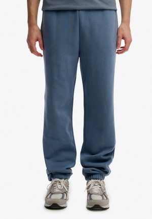 Person trägt blaue Jogginghosen mit elastischen Bündchen und graue Sportschuhe, steht vor einem schlichten Hintergrund.