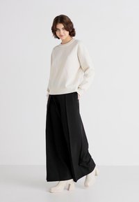 Krem genser, løs passform, rund hals; kombinert med svarte vide bukser; modellen har på seg hvite platåhæler. Glatt tekstur, minimalistisk design.