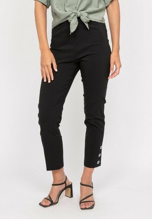Femme portant un pantalon slim noir taille haute avec trois boutons à la cheville, associé à des talons noirs à brides et un haut vert noué.