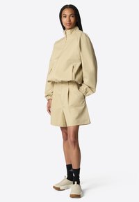 Chaqueta beige con un cuello alto y dos bolsillos con cremallera, combinada con pantalones cortos a juego. Usada con calcetines negros y zapatillas beige de tipo slip-on.