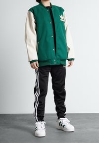 Vihreä varsity-takki, jossa on kermaan vivahtavat hihat ja Adidas-logo. Yhdistetty mustiin housuihin, joissa on valkoiset raidat, sekä valkoisiin lenkkareihin.