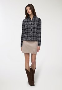 Camicia a quadri nera e grigia con maniche lunghe e due tasche sul petto, abbinata a una minigonna in suede beige con uno spacco laterale e stivali da cowboy marroni.