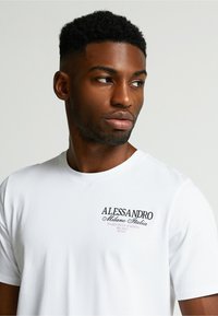 Biele bavlnené tričko s čiernym a fialovým potlačeným logom na ľavej hrudi, okrúhlym výstrihom a krátkymi rukávmi. Jednoduchý dizajn.