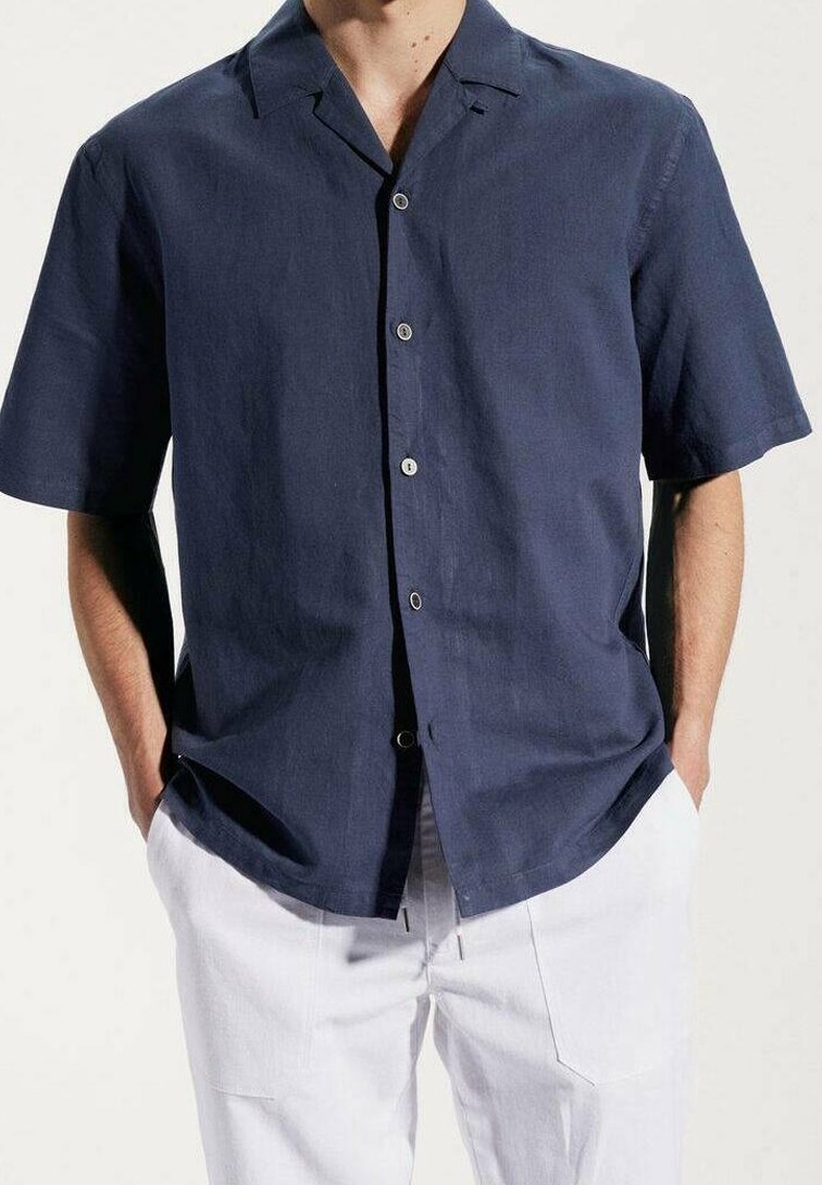 Granatowa koszula z krótkim rękawem wykonana z lekkiego materiału, z kołnierzem z wycięciem, przednimi guzikami i luźnym krojem. Noszona z białymi spodniami.