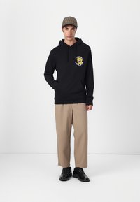 Felpa nera con cappuccio, caratterizzata da una grafica di un girasole e testo sul petto, abbinata a pantaloni larghi beige e scarpe nere.
