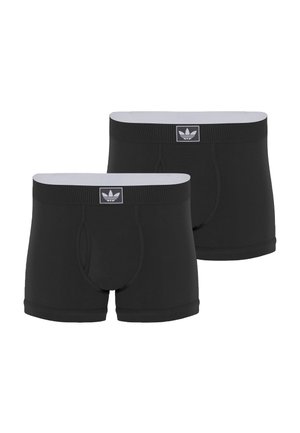Skjorteløs mand står barfodet, iført sorte Adidas boxershorts, justerer taljebåndet, vendt mod front på hvid baggrund.