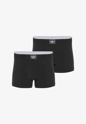 Ing nélküli férfi áll mezítláb, fekete Adidas boxeralsót visel, igazítja a derékpántot, előre néz egyszerű fehér háttér előtt.