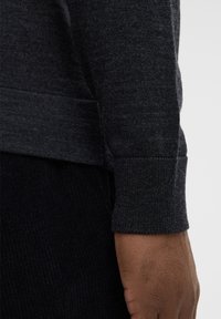 Dunkelgrauer Pullover mit strukturiertem Strick, eng anliegenden Bündchen und glatter Oberfläche am Körper. Eine sichtbare Hand ruht teilweise auf dem Bündchen.