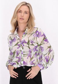 Bluză transparentă cu un design floral, având irisuri violete pe un fundal crem, închidere cu nasturi și mâneci lungi cu detalii la manșetă.