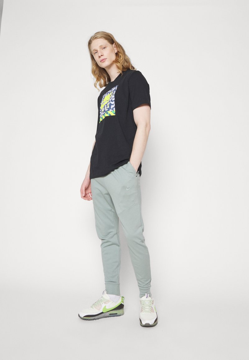 T-shirt noire avec un design graphique coloré, assortie à un jogger vert clair et des baskets blanches avec des accents verts, sur un fond blanc.