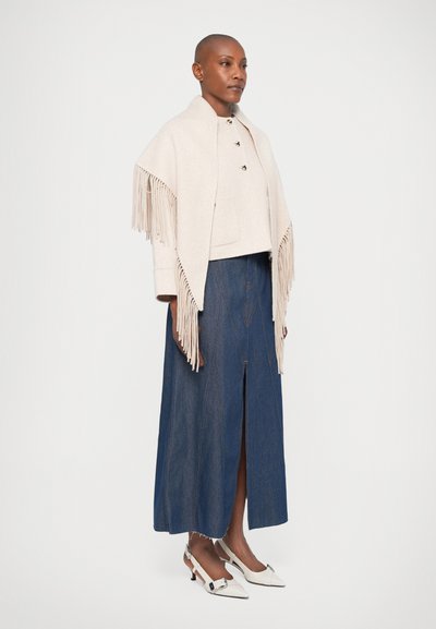 Beige fransad sjal över en kortärmad topp, kombinerad med en lång denimkjol och vita klackskor. Släta texturer, lång silhuett.
