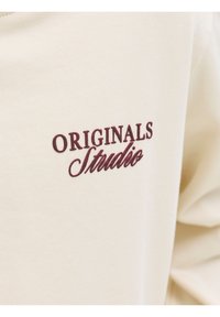 Κοντινό πλάνο μπεζ υφάσματος με μπορντό κεντημένο κείμενο «ORIGINALS Studio» σε ένα ένδυμα.