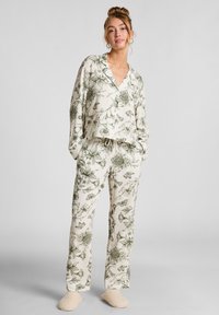 Vrouw in crèmekleurige pyjama met groene bloemen- en vlinderprint, staand met handen in de zakken, met beige slippers aan.