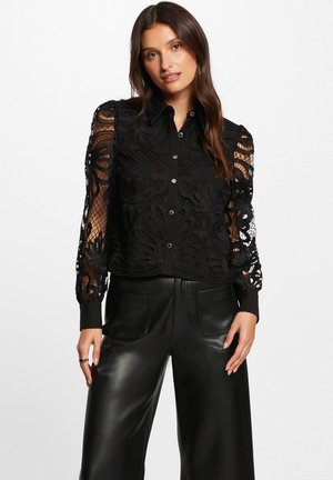Blusa di pizzo nero con motivi floreali intricati, dotata di una chiusura a bottoni frontale, maniche lunghe trasparenti e colletto nero solido. Indossata con pantaloni in pelle a vita alta.