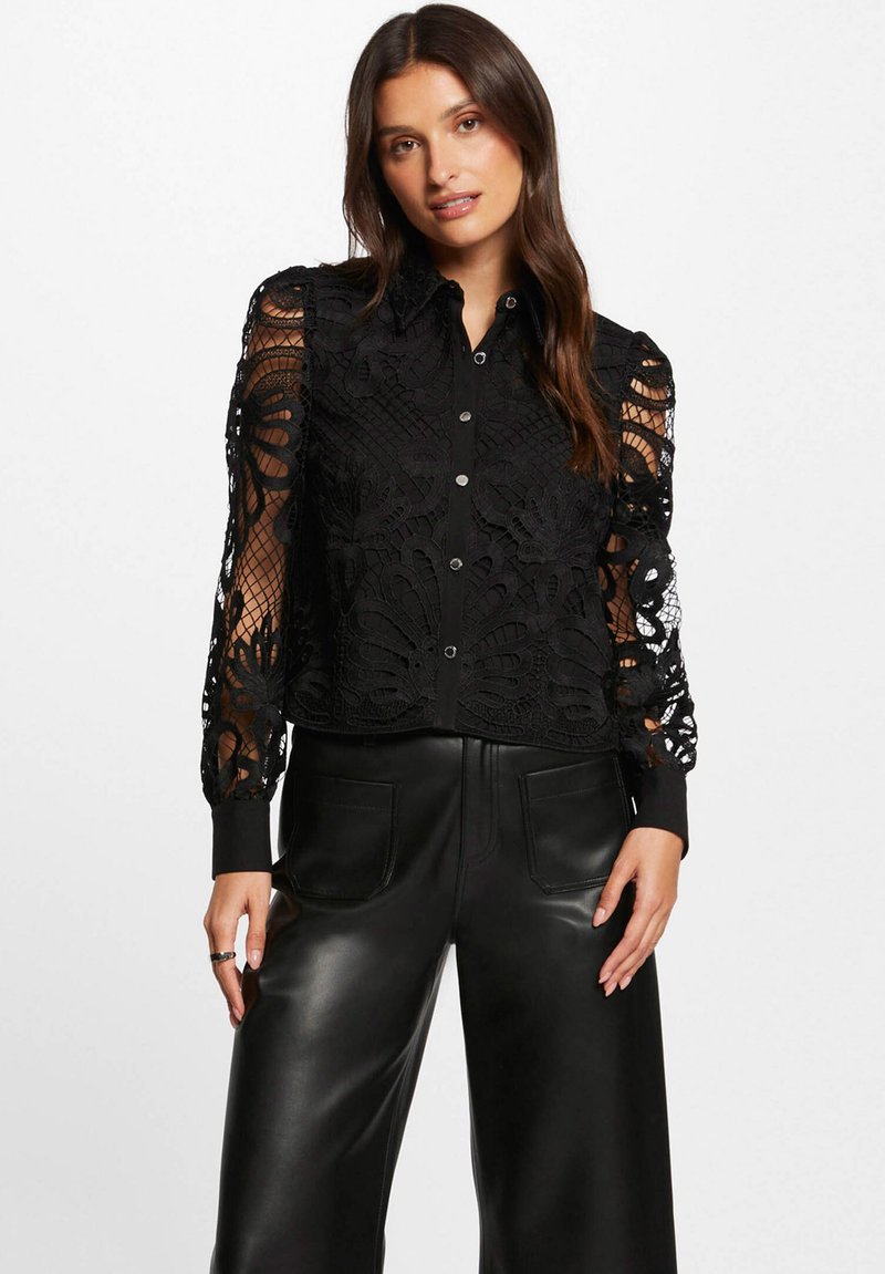 Blusa di pizzo nero con motivi floreali intricati, dotata di una chiusura a bottoni frontale, maniche lunghe trasparenti e colletto nero solido. Indossata con pantaloni in pelle a vita alta.