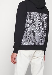 Sudadera negra de algodón con un gran patrón gráfico blanco y gris en la parte posterior. Presenta un bolsillo tipo canguro y puños acanalados.