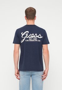 Hombre con una camiseta de Guess azul marino con el texto blanco "Guess EST. 1981 LOS ÁNGELES, CA" en la parte de atrás, combinada con jeans azul claro.