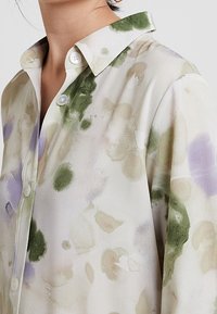Knapp-up skjorta med ett blommönster i grönt och lila på ett vitt strukturtyg, med krage och synliga knappar.