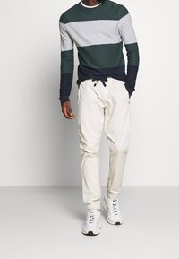 Pull rayé en vert foncé, gris et bleu marine, col rond. Jogging clair à taille élastique avec poches. Chaussures de sport blanches.
