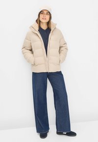 Beige pufferjakke med en krave i pels, lynlås-lukning og store quiltede sektioner. Kombineret med brede blå denimbukser og en lys kasket.