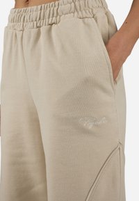 Beige joggebukser laget av myk bomullsblanding, med elastisk midjebånd, sidelommer og broderte logo detaljer på beinet.