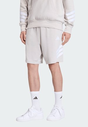 Persona che indossa pantaloncini beige chiaro e felpa abbinata con strisce bianche, calzini bianchi e sneakers bianche con loghi neri.