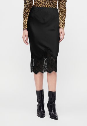 Vrouw draagt een zwarte satijnen midi-rok met kanten zoom, een leopardprint top met lange mouwen en glanzende zwarte enkellaarzen, staand op een effen achtergrond.