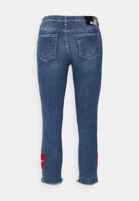 Vaqueros de denim azul con corte ajustado, que cuentan con dos bolsillos traseros, un dobladillo deshilachado y un detalle de corazón rojo en la rodilla izquierda. Logo negro en la cinturilla.