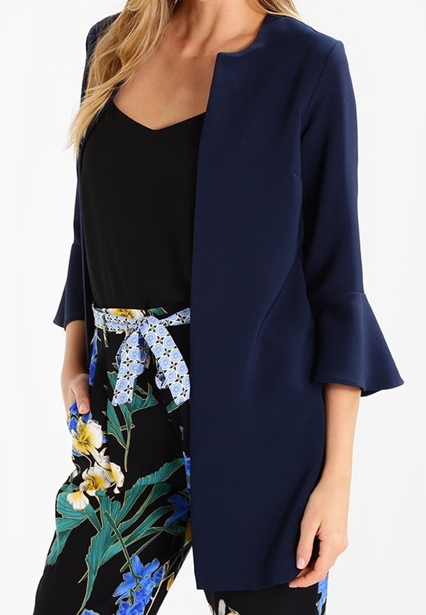 Marinblå öppen jacka med trekvartslånga trumpetärm, tillsammans med en svart topp och blommönstrade byxor med ett ljusblått bälte.