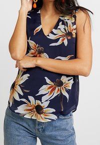 Top senza maniche blu navy con scollatura a V, caratterizzato da grandi motivi floreali gialli e bianchi su un tessuto leggero. Abbinato a jeans in denim blu.