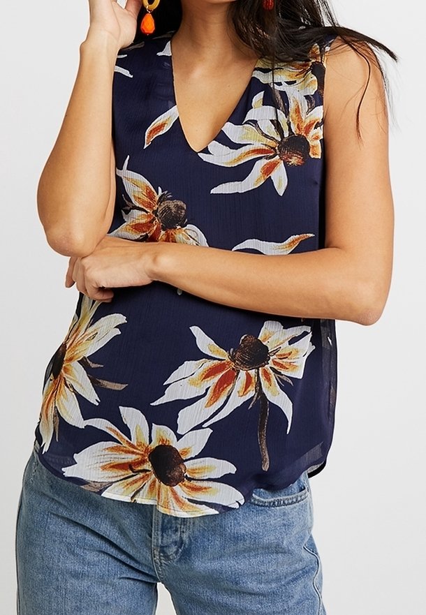 Top senza maniche blu navy con scollatura a V, caratterizzato da grandi motivi floreali gialli e bianchi su un tessuto leggero. Abbinato a jeans in denim blu.