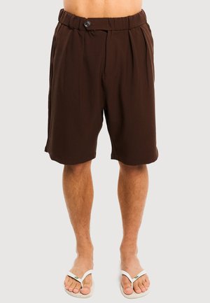 Barfodet person iført mørkebrune knælange shorts med knaplukning i taljen og hvide flipflops på en lys baggrund.