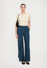 Beige přiléhavý top, široké modré kalhoty s drapovaným předním dílem, šikmé kapsy a černé boty na podpatku. Modelka nese malou černou kabelku.