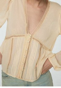 Blusa gialla pallido con scollatura a V profonda, texture plissettata, dettagli in pizzo e chiusura con bottoni sul davanti. Tessuto leggero e fluido.