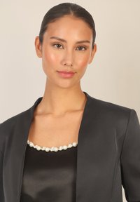 Mustaksi bleiseri, jossa on tyylikäs muotoilu ja satiinipinta, päällä musta satiinipaita, jossa on helmikaulakoru kaula-aukossa.