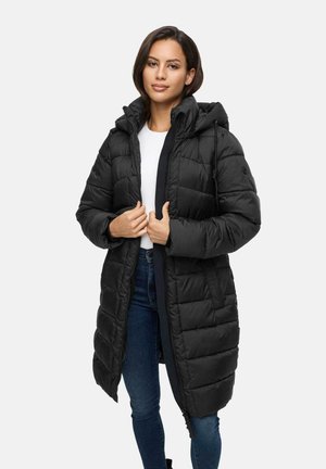 Woman wearing a long black quilted puffer coat with a hood, white shirt, and blue jeans, standing against a plain white background.
